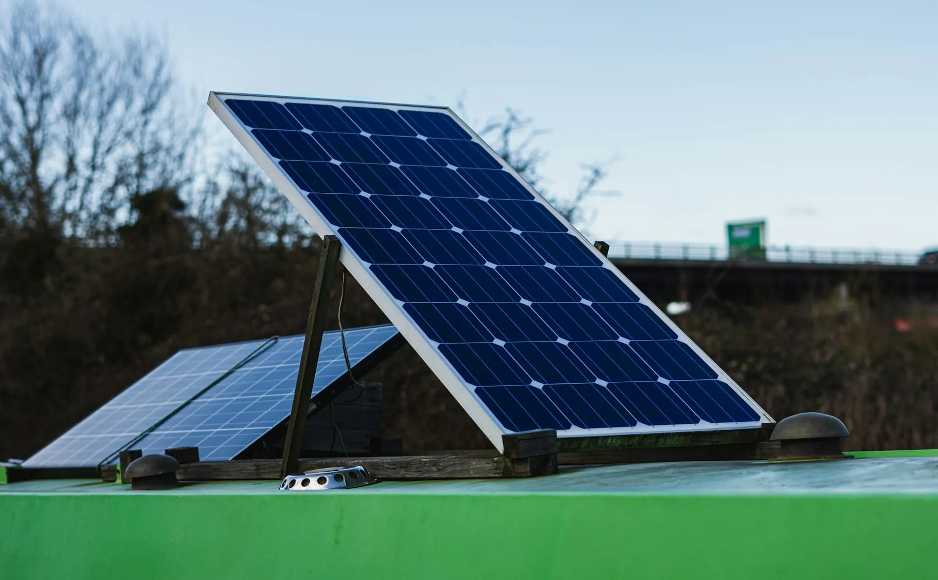 Two solar panels propped up on the green roof of a van beside a motorway bridge
