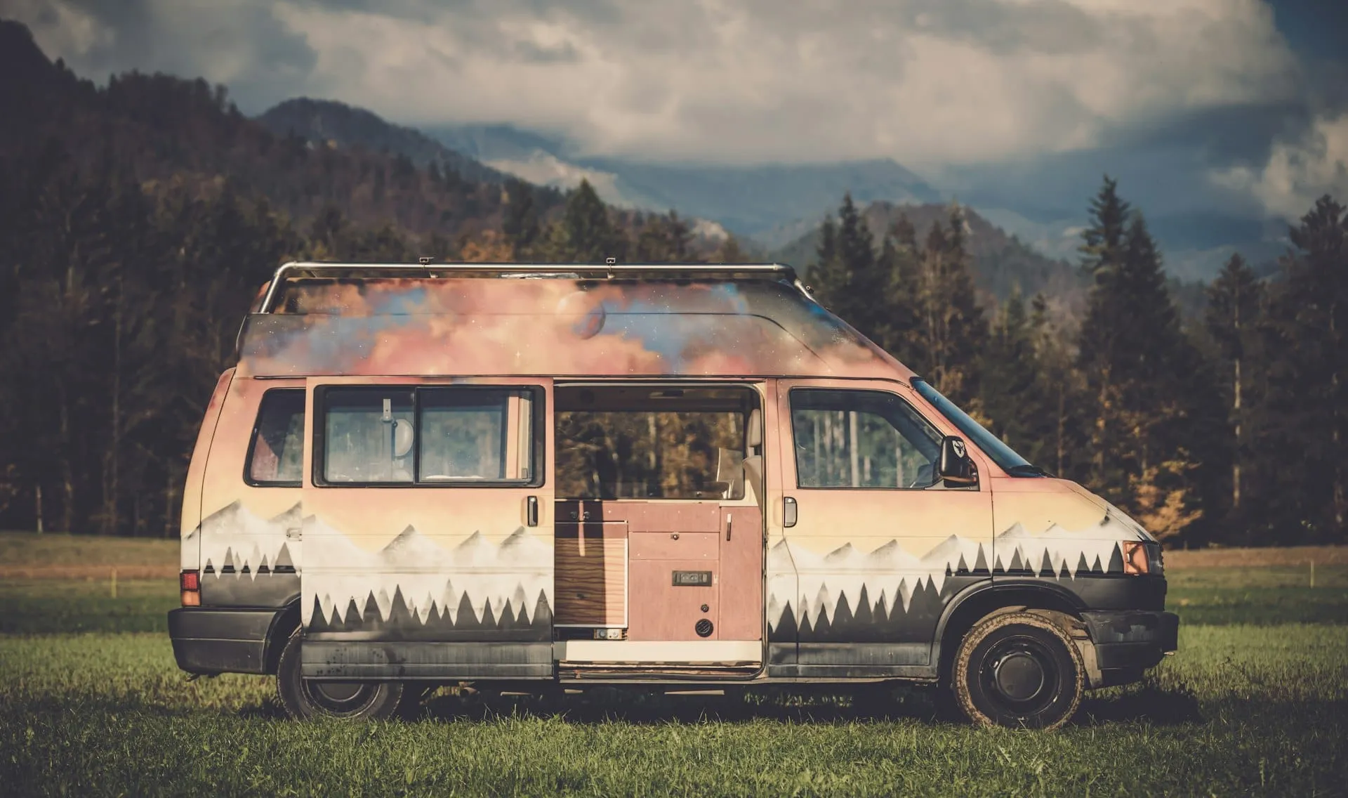 A painted campervan parked on grass in front of forested mountains with its sliding door open onto a small kitchen unit